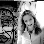 White woman with white blouse leaning against wall. Large Graffiti of a mask on the wall at the back with the word "Resist!: on the forehead.