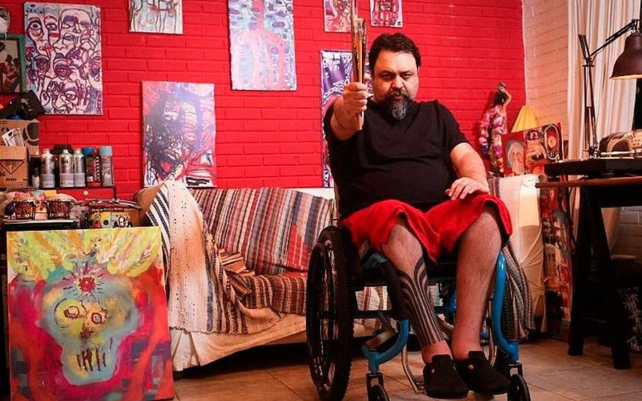 Marcelo Yuka - a masculine person, dark hair, sitting in a wheelchair and holding paintbrushes, surrounded by colourful artwork, on a red brickwall.