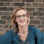 Caucasian middle-aged woman, fair hair, teal cotton blouse, a brick wall in the background.