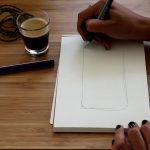 table, note pad, two hands, dark skin tone, one holding a pen and drawing on the notepad , glass filled with coffee and a pen on the table